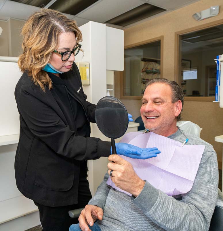 A female staff member with a male patient in the exam room looking into a mirror while smiling at Family Dental Practice in Kokomo IN.